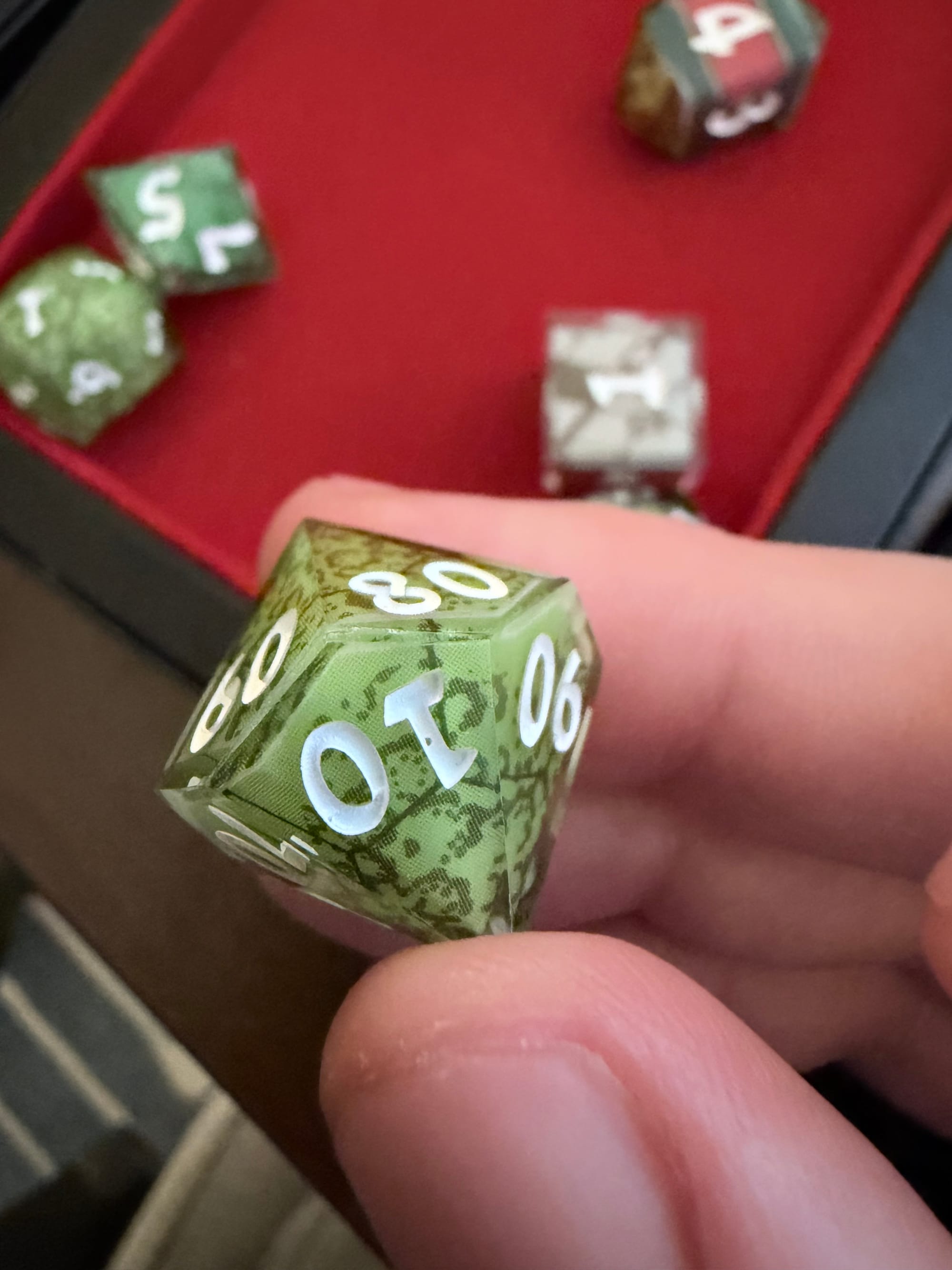 two white fingers holding a 10-sided die, with the "10" facing the viewer but upside down. other dice are on a red felt background, out of focus.